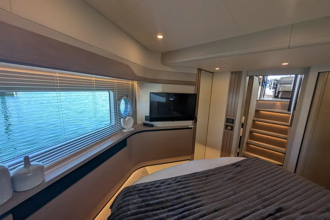 Modern yacht interior featuring a bed, television, round window, and stairs leading to upper deck. Natural light streaming in.